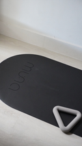 MUNA Oval Yoga Mat in black with non-slip performance and a light gray yoga accessory on a wooden floor.